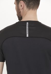 Sort atletisk T-shirt i fugttransporterende stof, med struktureret mønster, korte ærmer og et reflekterende "ENDURANCE"-label på bagsiden.
