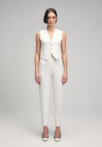 Witte op maat gemaakte gilet met een V-hals en knopen aan de voorkant, gecombineerd met slim-fit witte broek. Stiletto-hakken geven contrast met zwarte accenten.