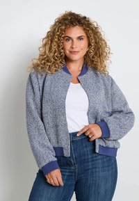 Femme avec des cheveux blonds bouclés portant une veste bomber texturée bleue, une chemise blanche et un jean bleu, se tenant devant un fond uni.