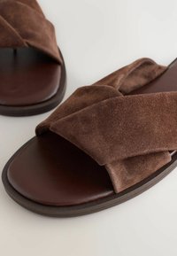 Bruine suède sandalen hebben een brede kruisriemontwerp, een ronde neus en een gladde donkerbruine zool, waarbij de zachte textuur en minimalistische stijl worden benadrukt.