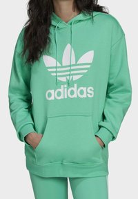 Grön huvtröja med en frontficka, dragsko i huvan och vitt Adidas-logotyp som har tre ränder och bladdesign. Mjuk tygstruktur.