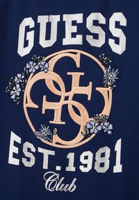 Tumesinine kangast T-särk, millel on virsiku- ja valge värvi tikandiga logo, koos lillemotiividega. Tekst sisaldab "GUESS EST. 1981 Klubi."