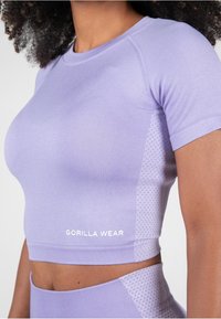 Violets krāsas cropped sporta tops ar ribotu tekstūru, ar sānu paneļiem, kas rotāti ar punktu rakstu, un „GORILLA WEAR” logo izdrukātu baltā krāsā.