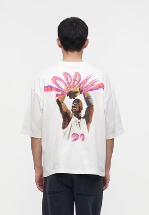 Homme portant un t-shirt blanc avec un graphisme d’un joueur de basketball en maillot blanc numéro 23 en train de shooter un ballon, le mot « JORDAN » au-dessus en lettres colorées.
