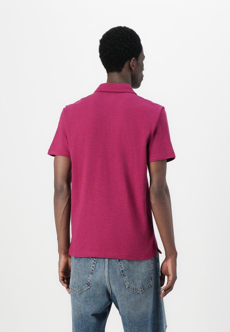 Polo à manches courtes de couleur rose foncé, fabriqué dans un tissu texturé. Doté d'un col classique et d'un ourlet droit avec de légentes fentes latérales.