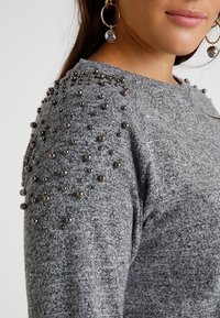 Grijze gebreide trui met een gestructureerde stof en versierde schouders met metalen kralen, wat zorgt voor een uniek designelement.