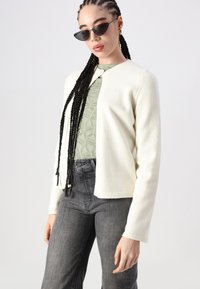 Cardigan de malha em tecido suave creme, com decote redondo e frente aberta, combinado com um top texturizado verde claro e calças cinza-escuras.