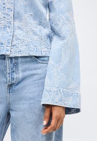 Ανοιχτό μπλε τζιν μπουφάν και τζιν παντελόνι με λευκή κέντηση με λουλούδια, εστίαση στο μανίκι, την τσέπη και το χέρι με ανοιχτόχρωμο δέρμα και λευκό βερνίκι νυχιών.