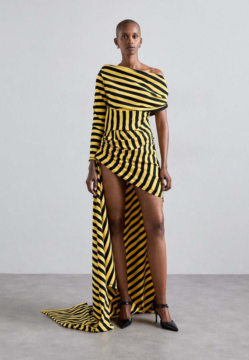 Gul-och svartrandig off-shoulder klänning med asymmetrisk fåll, veckad design och ett långt släp. Buren med svarta högklackade skor.