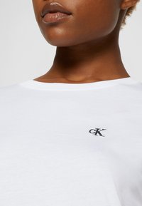 Vit bomullströja med rund halsringning som har en liten svart "cK"-logotyp broderad på vänster bröstområde. Närliggande textur.