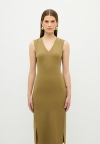 Olijfgroene, aansluitende, mouwloze jurk met een V-hals, zijsplijten en minimale naadtoeslagen. Glad, sleek stoftextuur. Gouden armband.