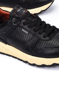 Zapatilla negra de cuero con laterales texturizados, cordones redondeados y una suela de goma color crema. Presenta una etiqueta con el logotipo y costuras decorativas.