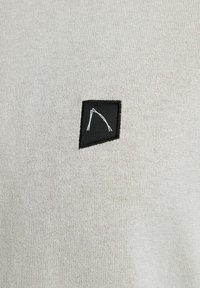 Lichtgrijze gebreide pullover met een gestructureerd oppervlak. Bevat een klein zwart label met een witte abstracte afbeelding in de linker bovenhoek.