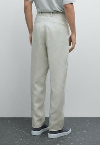 Pantaloni beige chiaro con una texture liscia, caratterizzati da una vestibilità su misura, tasche laterali e tasche sul retro a filetto. Indossati con sneakers grigie.