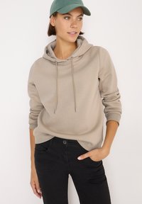 Beige mikina s kapucí a stahovací šňůrkou se stříbrnými koncovkami, vybavená klokaní kapsou. Nosená s černými džíny a zelenou čepicí. Hladký povrch.