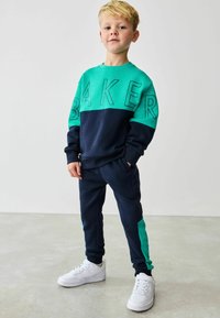 To-delt drenge træningssæt med en turkis top med "BAKER" i fede bogstaver, parret med marineblå sweatpants og hvide sneakers.