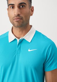 Turkusowa koszulka polo Nike z białym kołnierzykiem i małym logo na piersi. Materiał ma teksturowany, żebrowany wygląd.