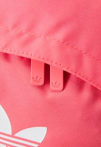 Ryškiai rožinė medžiaginė maišas su tekstūruota paviršiaus. Jame yra du rožinės spalvos užtrauktuko traukimai su reljefiniu logotipu ir didelis baltas logotipas vienoje pusėje.