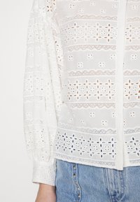 Blusa bianca con motivo in pizzo ricamato, maniche lunghe e voluminose, e chiusura con bottoni. Abbinata a jeans in denim azzurro chiaro.
