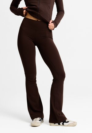 Kvinde iført mørkebrune tætsiddende vide bukser og matchende jakke med lynlås, løfter jakken for at afsløre maven, hvide sneakers synlige.