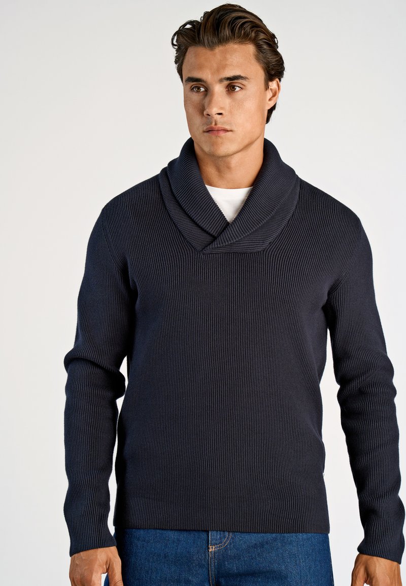 Marineblauer Rippstrickpullover mit umgeschlagenem Schalkragen, langen Ärmeln und lockerer Passform, kombiniert mit blauen Jeans, vor einem einfarbigen Hintergrund.