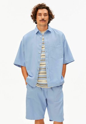 Mann mit lockigem Haar und Schnurrbart, der ein hellblaues, kurzärmeliges Hemd mit Knöpfen und passende Shorts über einem gestreiften T-Shirt trägt, stehend mit den Händen in den Taschen.