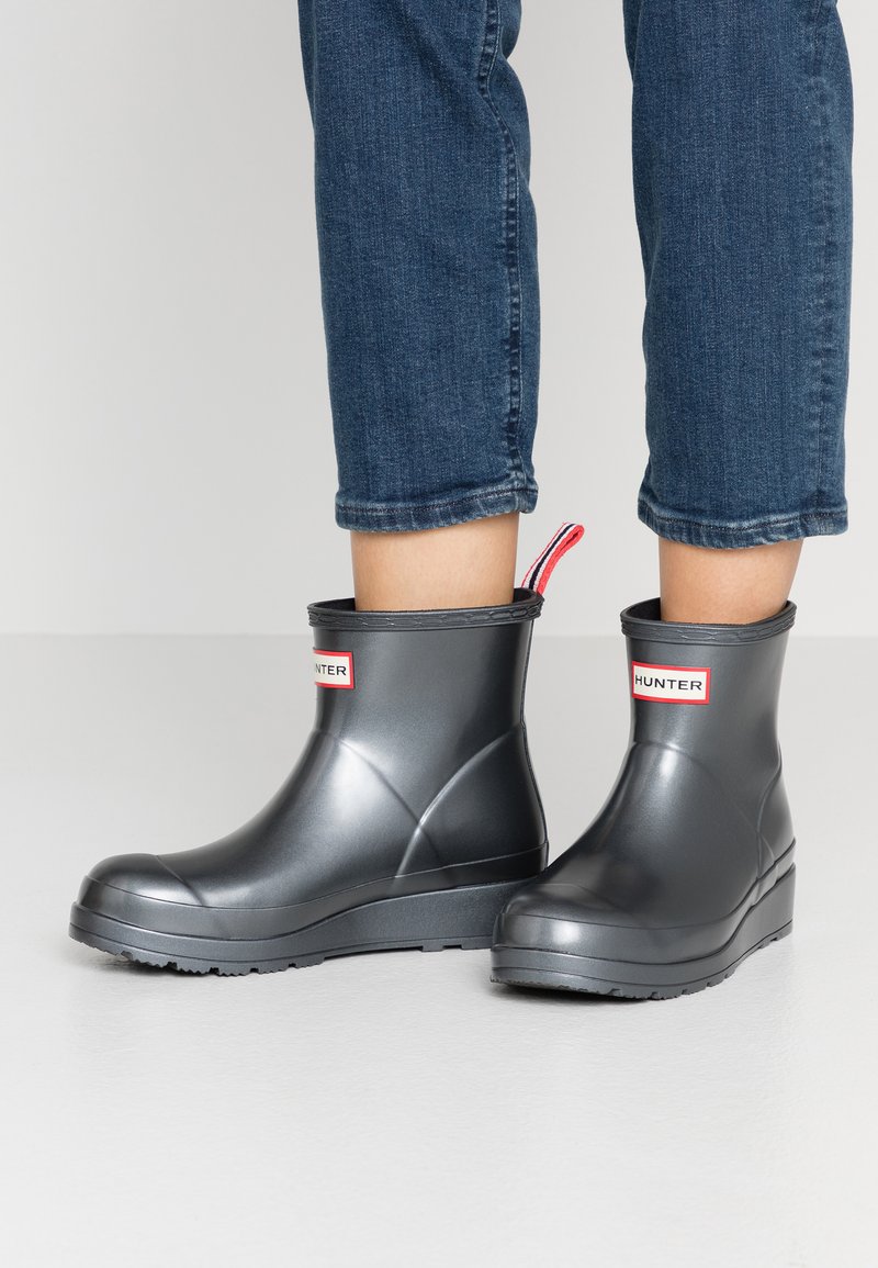 Botas de goma gris a la altura del tobillo con punta redonda y suela gruesa, que cuentan con una etiqueta de logo en rojo y blanco en la parte delantera y una lengüeta con rayas.