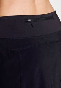 Cintura atlética negra fabricada con un tejido liso y elástico. Presenta un pequeño bolsillo con cremallera y costuras de contraste en la parte superior.