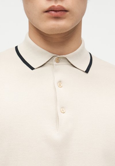 Άνδρας που φοράει μπεζ πουκάμισο πόλο με γιακά με μαύρο περίγραμμα και τρία κρεμ κουμπιά κουμπωμένα στο λαιμό.