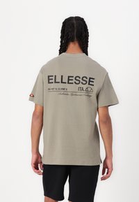 Camiseta de algodón gris con mangas cortas, que presenta un gran logo negro de "ELLESSE" y coordenadas impresas en la parte posterior; dobladillo plano, corte relajado.