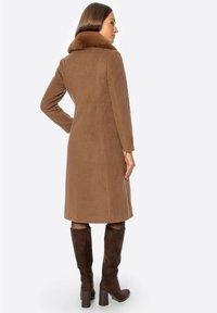Brązowy płaszcz z mieszanki wełny z miękkim kołnierzem, długimi rękawami i prostym krojem, noszony z wysokimi brązowymi zamszowymi kozakami, widziany od tyłu.