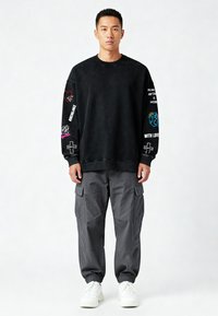 Črna puhasta majica s kapuco z večbarvnimi grafičnimi motivi na rokavih, v kombinaciji s sivimi cargo hlačami z stranskimi žepi in belimi supergami.