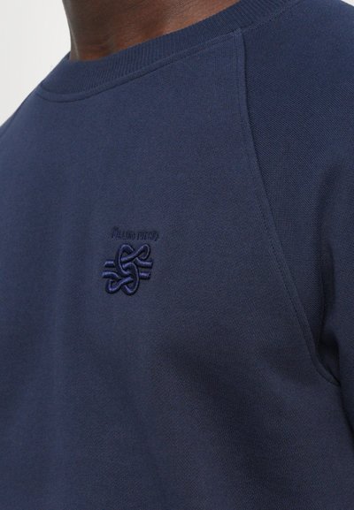 Tuvumā redzams tumši zils krekls ar izceltiem izšūtiem mezglu logotipu un "Filling Pieces" tekstu uz krūšu daļas.