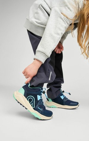 Dziecko poprawiające mankiet ciemnych spodni, noszące granatowo-miętowe trampki z białymi podeszwami na jasnoszarym tle.