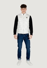 Hvid polstret vest med lynlås foran, sort langærmet skjorte under, blå jeans og hvide sneakers. Glat tekstur med et quiltet mønster.