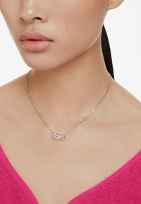 Collier en chaîne argentée avec un pendentif en forme d'infini rose décoré de strass, sur fond uni.