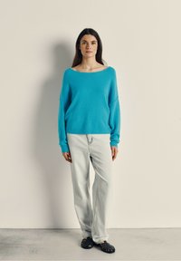 Blå, oversized sweater med en bred udskæring og lange ærmer; parret med lyse denimjeans og sorte vævede sandaler. Neutral baggrund.
