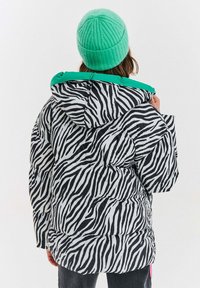 Černobílá bunda Puff s pruhy jako na zebře a zelenou kapucí. Má volný střih a prošívanou texturu, pohled zezadu.