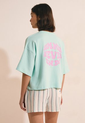 Młoda kobieta z ciemnymi włosami do ramion, ubrana w jasnoniebieski, oversize'owy T-shirt z różowym napisem "SUMMER NEVER ENDS" oraz krótkie spodenki w paski.