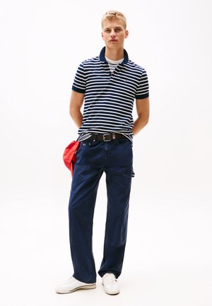Polo cu dungi bleumarin, cu mâneci scurte, purtat cu pantaloni largi bleumarin. Adidași albi și un accesoriu roșu completează ținuta.
