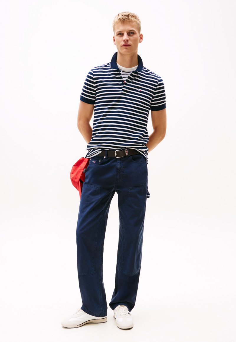 Polo à rayures marines à manches courtes, associé à un pantalon ample marine. Des sneakers blanches et un accessoire rouge complètent la tenue.