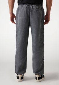 Pantalones grises, holgados, con cinturilla elástica y bolsillo trasero. Textura suave con un aspecto ligeramente arrugado, combinados con zapatos negros.