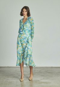 Abito con stampa floreale e orlo arricciato, nelle tonalità di blu, verde e giallo. La parte superiore presenta un profondo scollo a V e maniche lunghe.