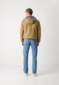 Khakifarbene Kapuzenjacke mit glatter Textur, isolierender Füllung und elastischen Bündchen, kombiniert mit hellblauen, geraden Jeans und beigen Sneakers.