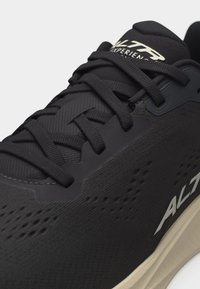 Κοντινό πλάνο μαύρου αθλητικού παπουτσιού με μαύρα κορδόνια, διάτρητο πλαϊνό πάνελ και μπεζ σόλα με την ένδειξη "ALTRA" στο πλάι και στη γλώσσα.