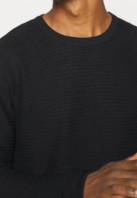 Man wearing a black ribbed long-sleeve shirt, folded arms visible, dark skin tone, and close-up on torso and neck area.