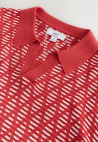 Polo en maille rouge avec des rayures horizontales blanches en pointillés et une étiquette de taille moyenne indiquant "NEXT Regular Fit M."