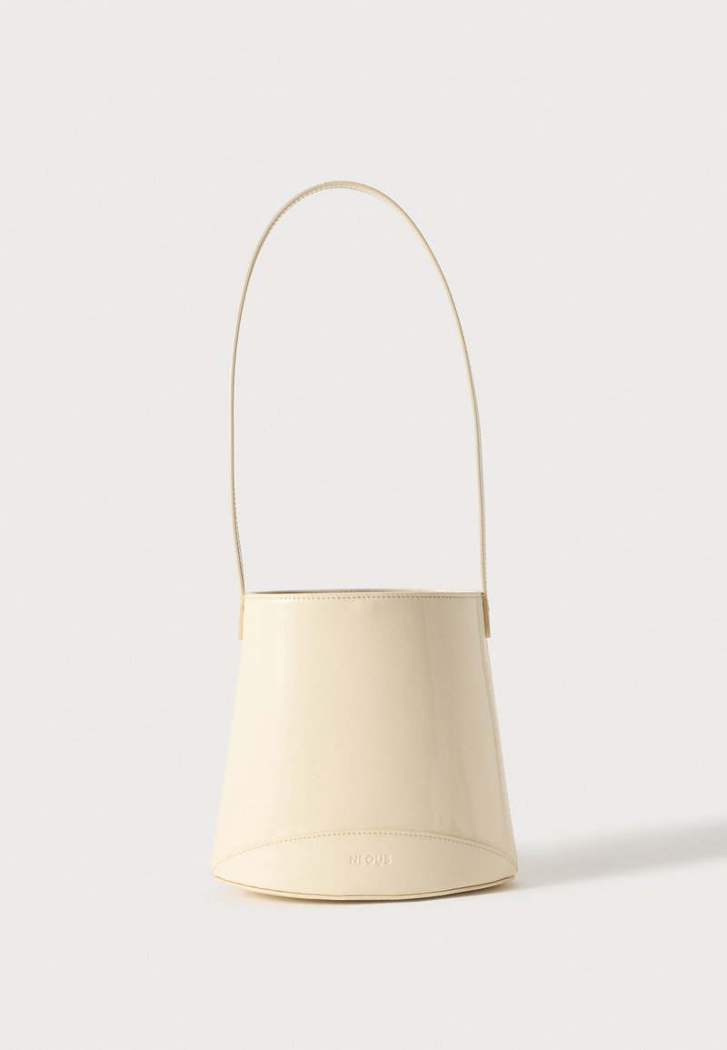 Sac seau en cuir couleur crème avec une longue sangle fine et des coutures discrètes, debout sur un fond uni.