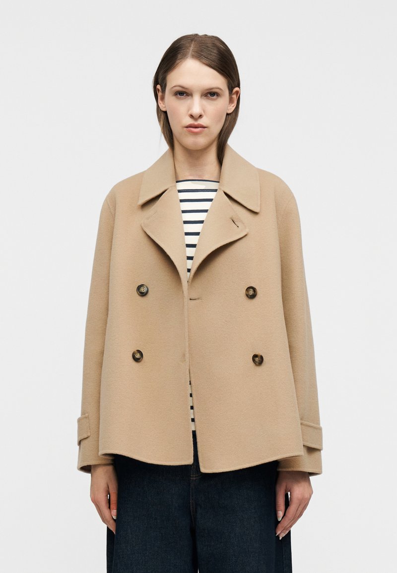 Γυναίκα με μπεζ διπλόπατο μάλλινο παλτό, ριγέ πουκάμισο και σκούρο τζιν με φαρδιά παντελόνια, στέκεται μπροστά σε απλό ανοιχτό φόντο.