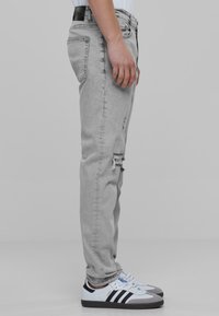 Hallid jeanid slim fit lõikega, millel on kulumise detailid põlve piirkonnas ja must silt vööl, kombineeritud valgete spordijalatsitega.